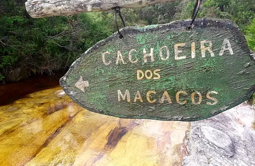 placa verde onde há escrito 'cachoeira dos macacos', ao fundo, piscina natural de cachoeira e árvores, durante o dia