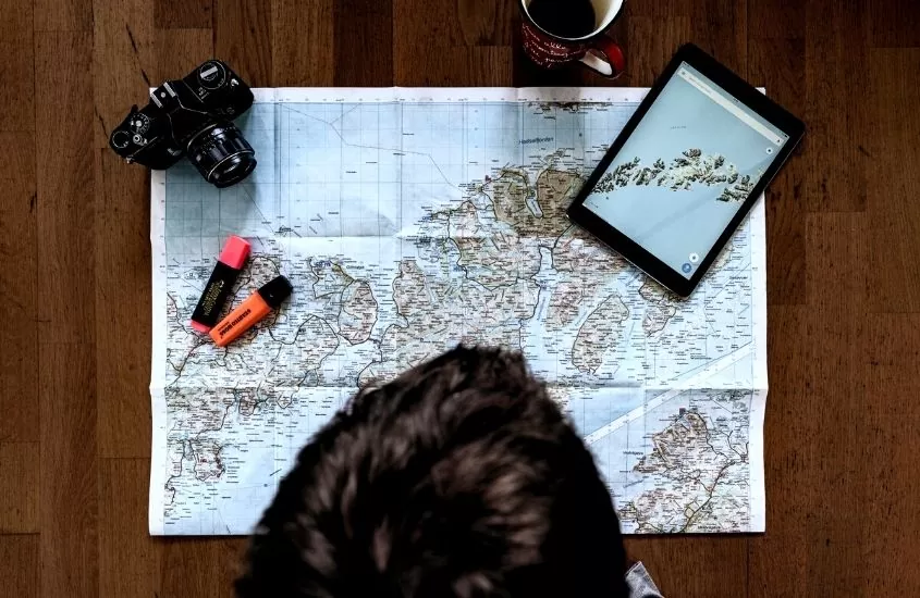 vista de cima de mesa onde há câmera, tablet, caneca e marcadores de texto em cima de um mapa de papel