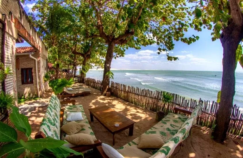 sofás, mesas e espreguiçadeiras cercadas de árvores em varanda com vista para o mar em Pousada Ponta Da Pedra, uma das pousadas em jericoacoara baratas