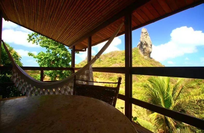 mesa, cadeira e rede de descanso em varanda com vista para as montanhas, durante o dia em fernando de noronha, em Pousada Zé Maria