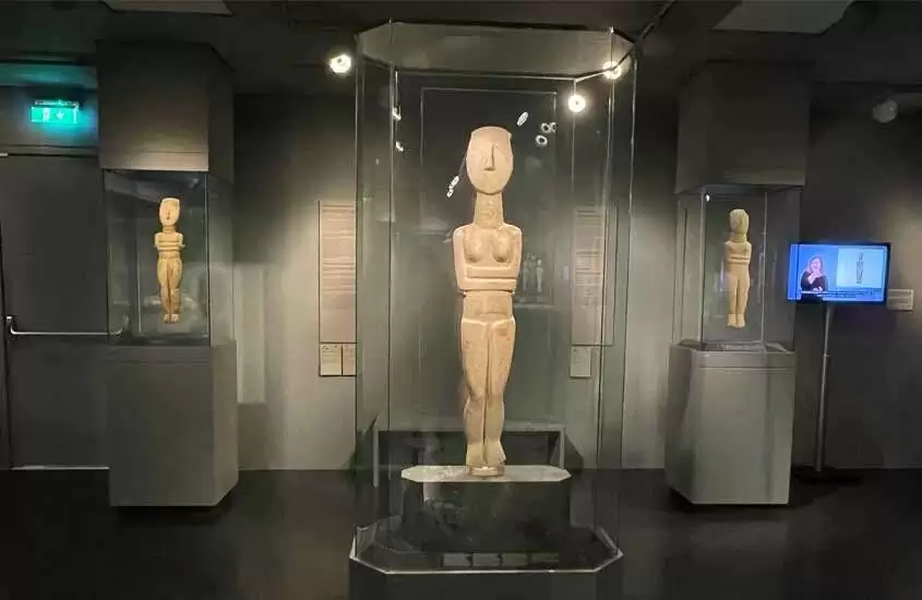 Interior de museu com estátuas em redomas de vidro e TV