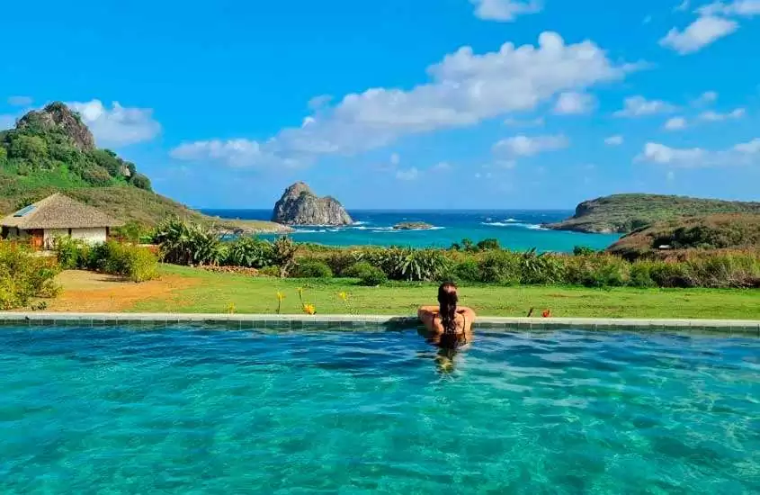 Em um dia de sol, piscina de borda infinita com 2 pessoas e vista para a praia e para as montanhas, ao lado animais silvestres e área gramada