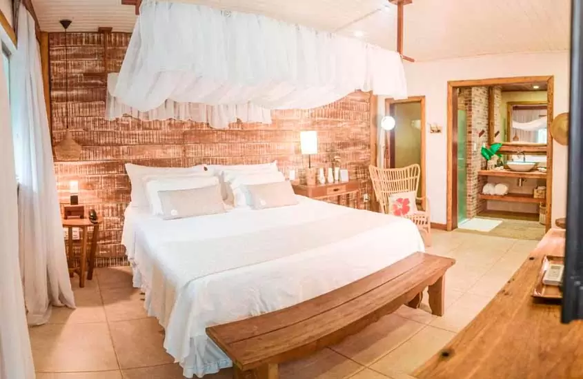 Quarto de uma das pousadas em Fernando de Noronha com cama de casal, móveis de madeira, luminárias, TV e banheiro do lado