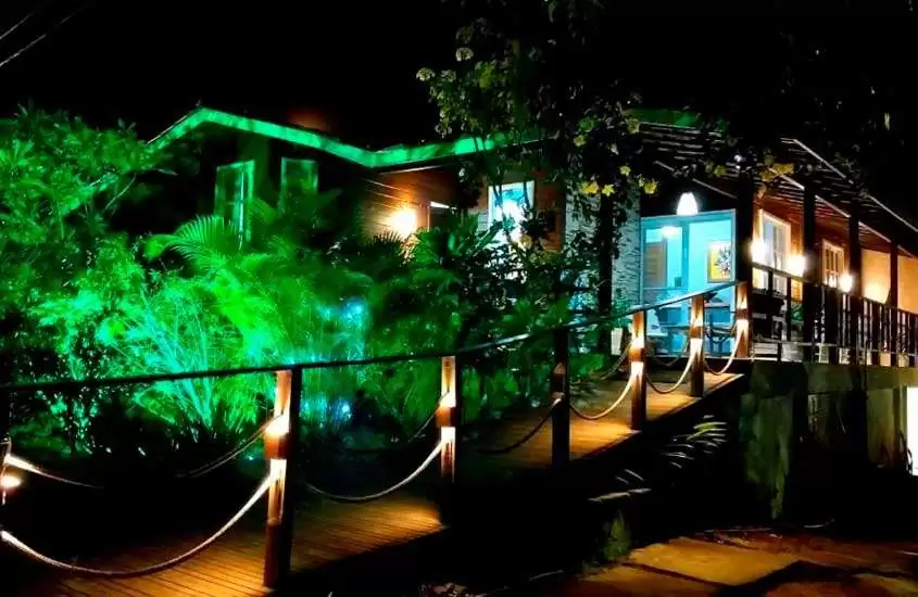 Durante a noite, fachada de uma pousada em Fernando de Noronha com ponte de madeira, plantas decorativas e luzes verdes iluminando