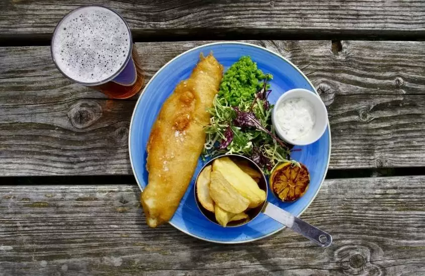 vista de cima de mesa de madeira onde há em cima copo de cerveja e prato azul com peixe, salada e batata
