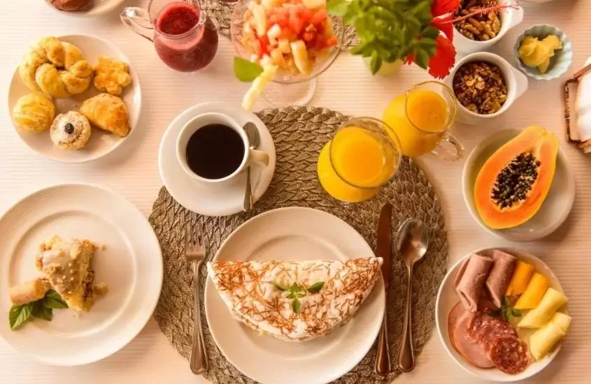 vista de cima de pães, sucos, café, tapioca em cima de mesa branca em café da manhã oferecido pela pousada corveta
