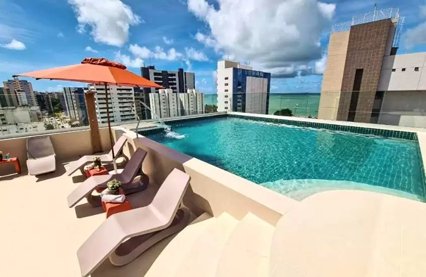 espreguiçadeiras e piscina com vista para prédios da cidade, durante dia ensolarado em área de lazer de Verano Pajuçara by Tropicalis