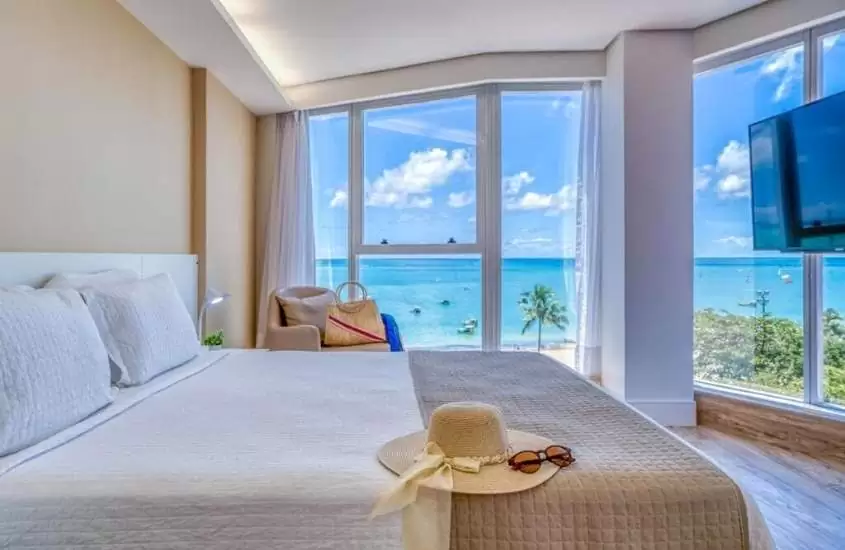 chapéu e óculos de sol em cima de cama de casal em suíte com ampla janela com vista para o mar, durante o dia, em Hotel Brisa Suites