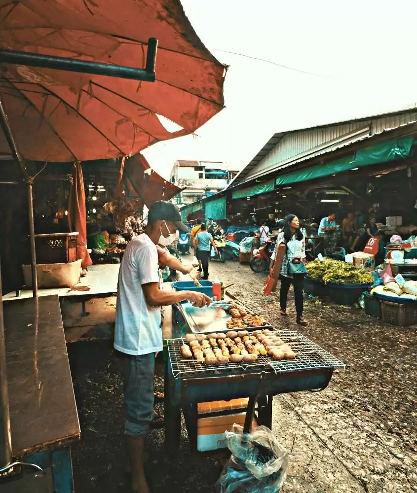 homem prepara churrasco em barraca de comida, em rua cheia de barracas da tailândia, durante o dia