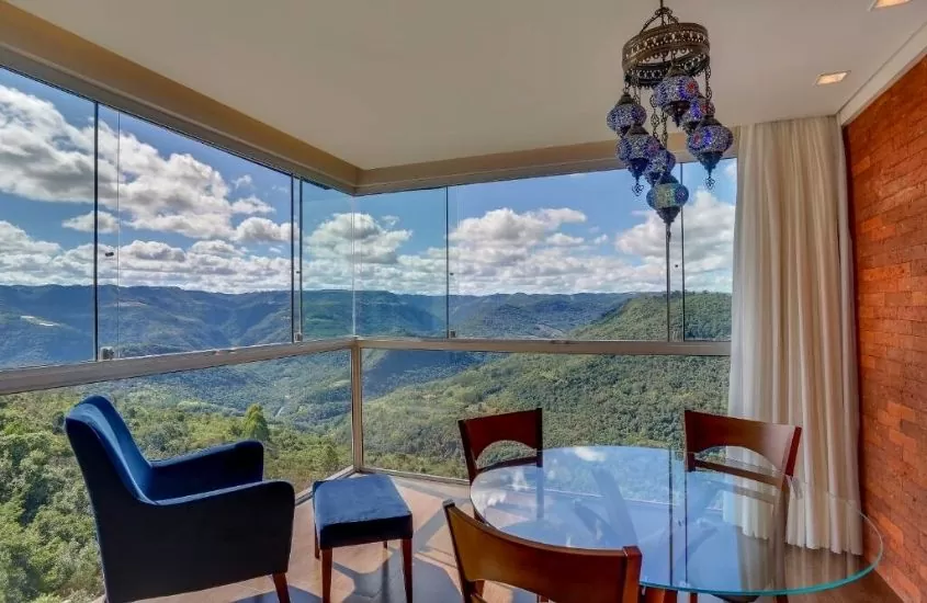 poltrona azul e cadeiras ao redor de mesa redonda em suíte com amplas janelas com vista para as montanhas, durante o dia em Pousada Janela do Vale