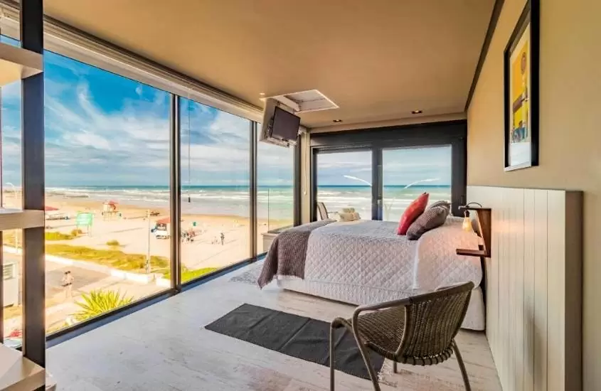 durante o dia, cama de casal e cadeira em suíte com amplas janelas com vista para a praia