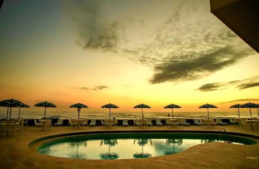 durante entardecer, espreguiçadeiras e guarda sóis ao redor de piscina com vista para o mar em área de lazer de Dunas Praia Hotel