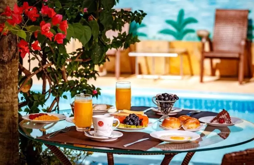 suco, frutas e pães em cima de mesa de café da manhã servido em frente a piscina em pousada Jardim dos Orixás