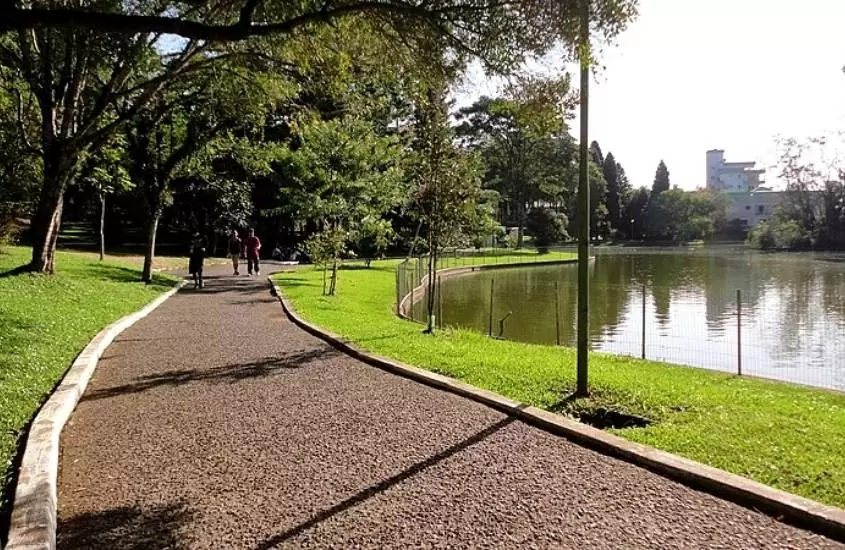 durante o dia, pessoas caminhando em ciclovia rodeada por árvores, em frente a lago em Parque dos Pinheiros, um dos pontos turísticos de farroupilha