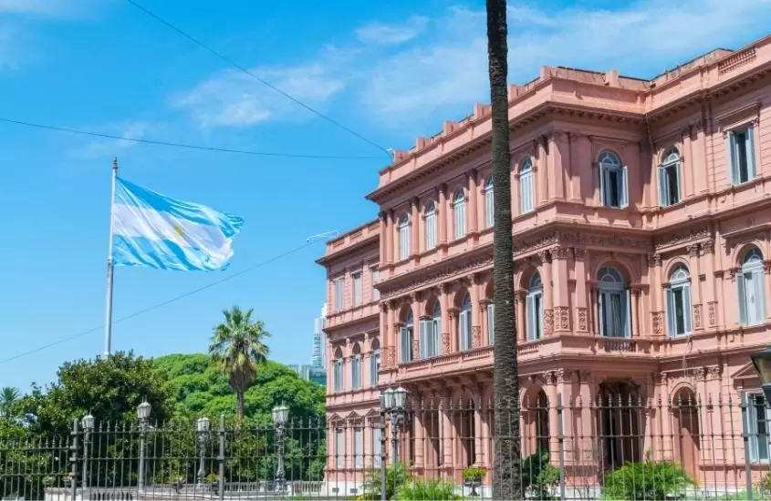durante o dia, bandeira da argentina hasteada em frente a construção rosa de três andares conhecida como casa rosada