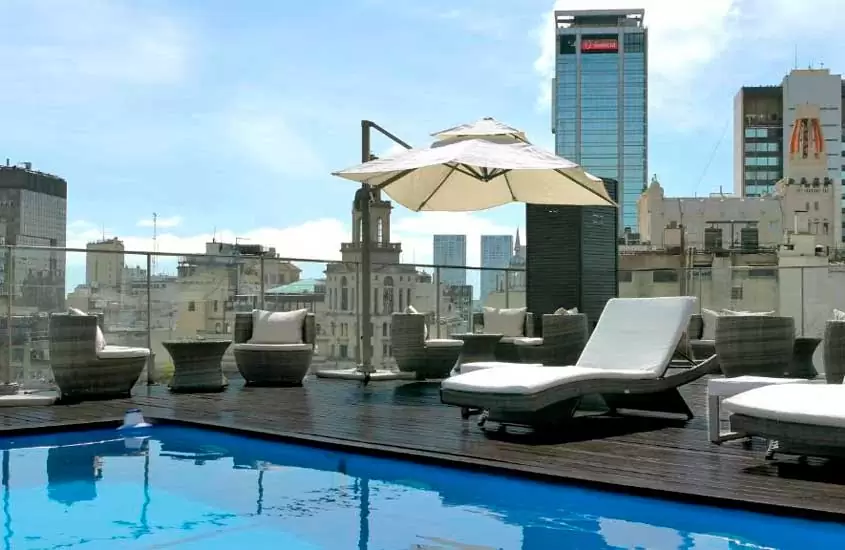 Em um dia de sol, área de lazer de um dos melhores hotéis em Buenos Aires com piscina, cadeiras, mesas, espreguiçadeira, ombrelone e vista da cidade ao redor