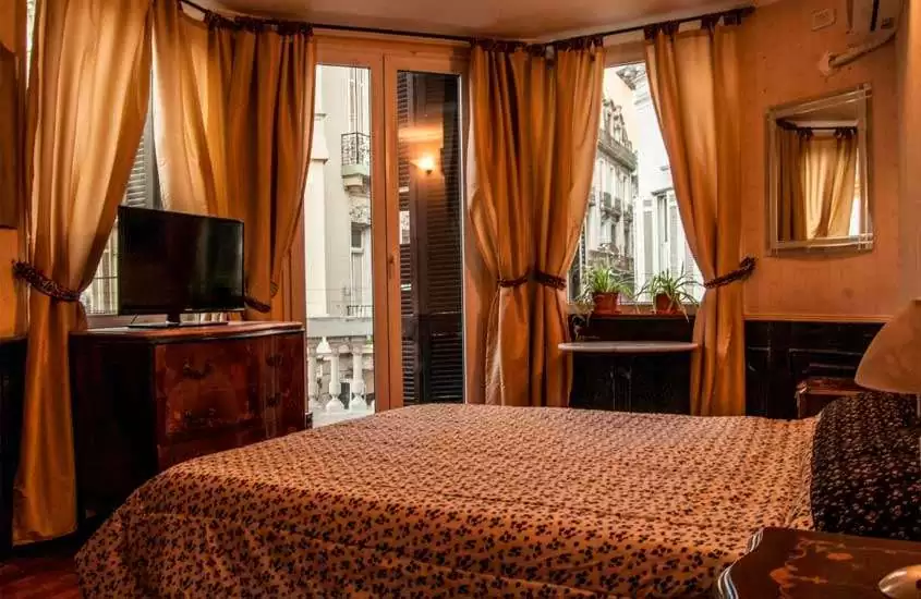 Quarto de um dos melhores hotéis em Buenos Aires com cama de casal, TV, cômoda de madeira, janelas grandes acortinadas, espelho e paisagem da cidade