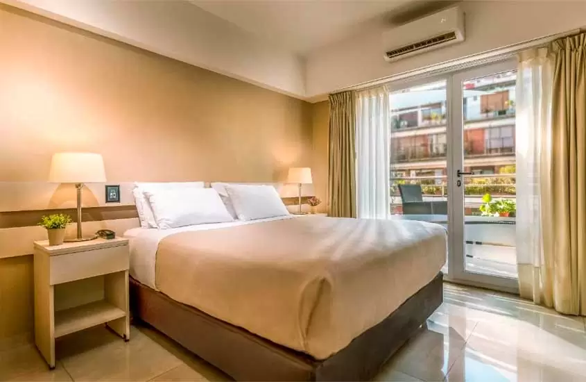 Quarto de hotel em Buenos Aires com cama de casal, comodar com luminárias, ar-condicionado e janela para varanda com poltrona