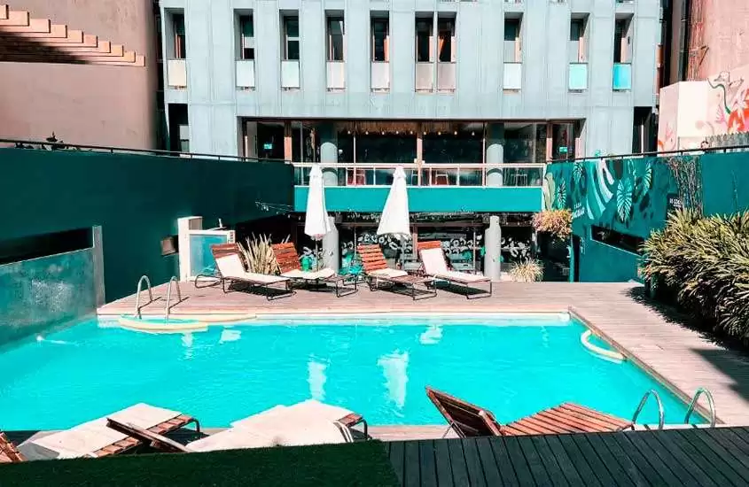 Em um dia de sol, área de lazer de hotel em Buenos Aires com piscina, espreguiçadeiras, guarda-sóis, deck de madeira e plantas ao redor