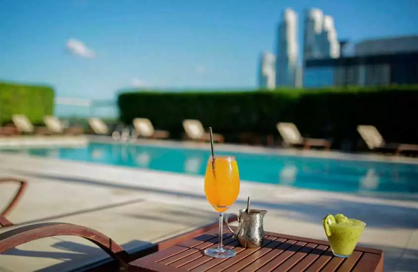 Em um dia de sol, área de lazer de hotel com piscina, espreguiçadeiras, paredes de grama e mesa de madeira com drinks