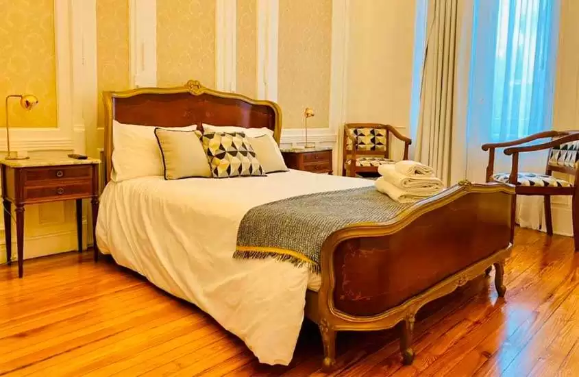 Quarto de hotel com cama de casal, poltronas, cômodas e janelas grandes acortinadas