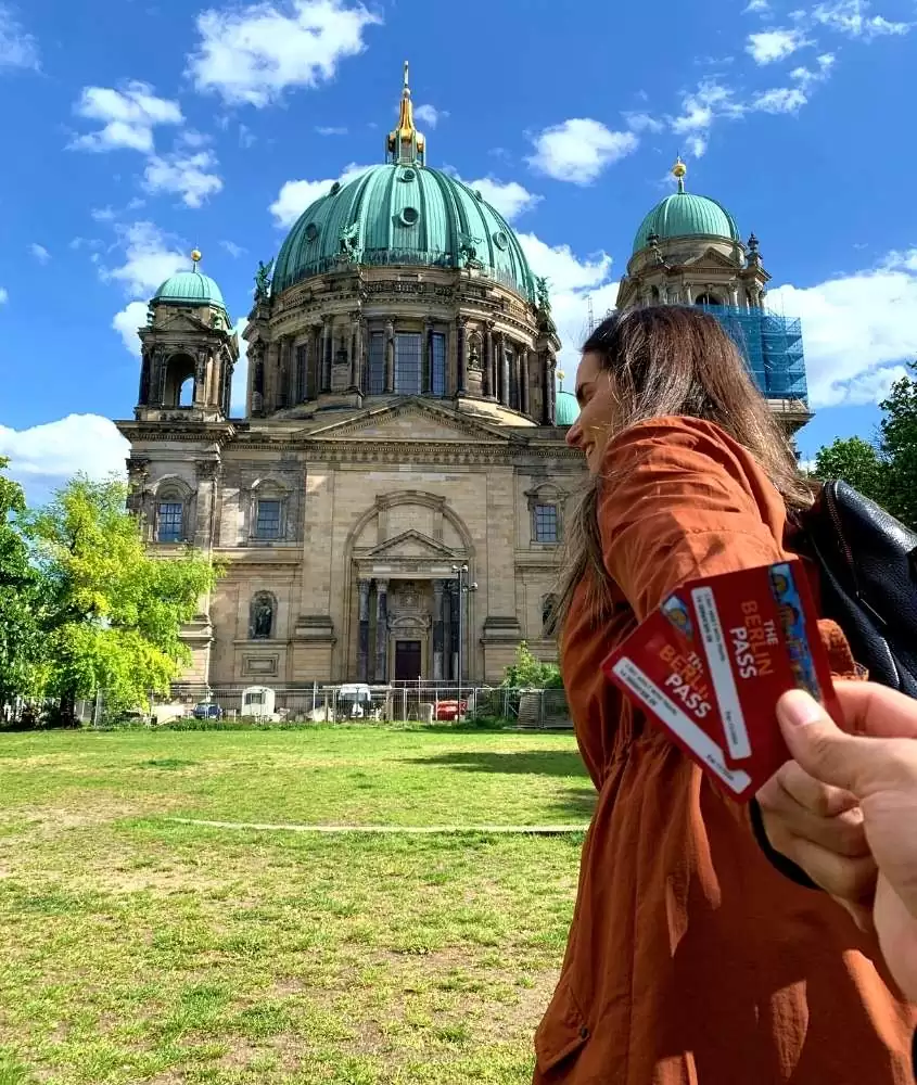 bárbara rocha com casaco laranja, sorri em frente a catedral de berlim, mostrando cartão do berlin pass, durante o dia