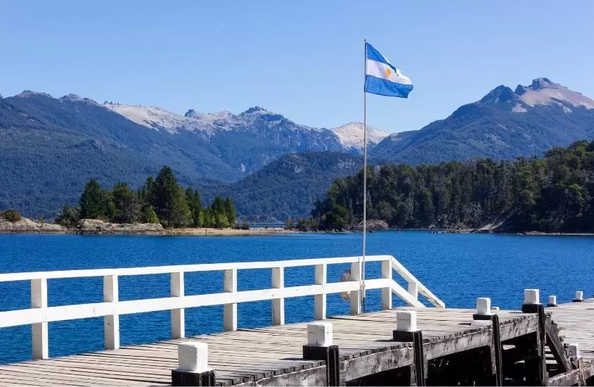bandeira da argentina hasteada em deck à beira de lago e montanhas ao fundo, durante o dia