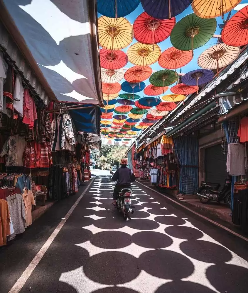 pessoa passando de moto, em rua coberta cheia lojas de roupa, sob teto cheio de guarda-chuvas coloridos