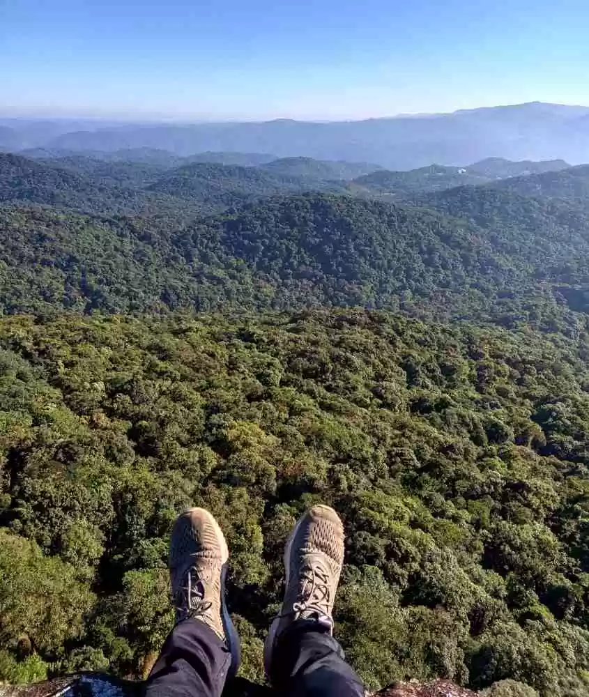durante o dia, pessoa com calça e tênis, com pernas esticadas em mirante com vista para as montanhas de Joinville