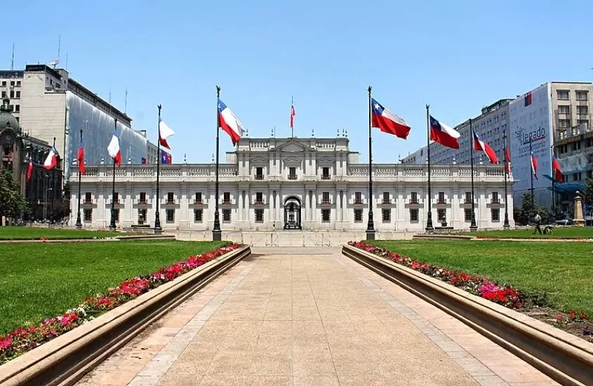 durante o dia, diversas bandeiras do chile hasteadas em frente a grande construção branca, onde funciona palacio de la moneda
