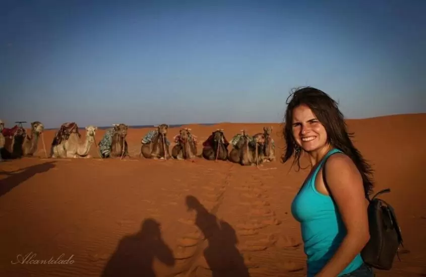 durante o dia, bárbara rocha, de regata azul, carregando mochila preta, sorri para foto; ao fundo, camelos sentados em areia do deserto do saara
