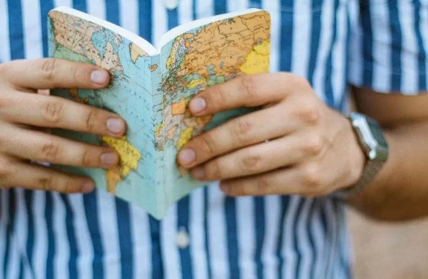 pessoa com camisa listrada branca e azul, segurando com as duas mãos, caderno azul com capa de mapa mundi