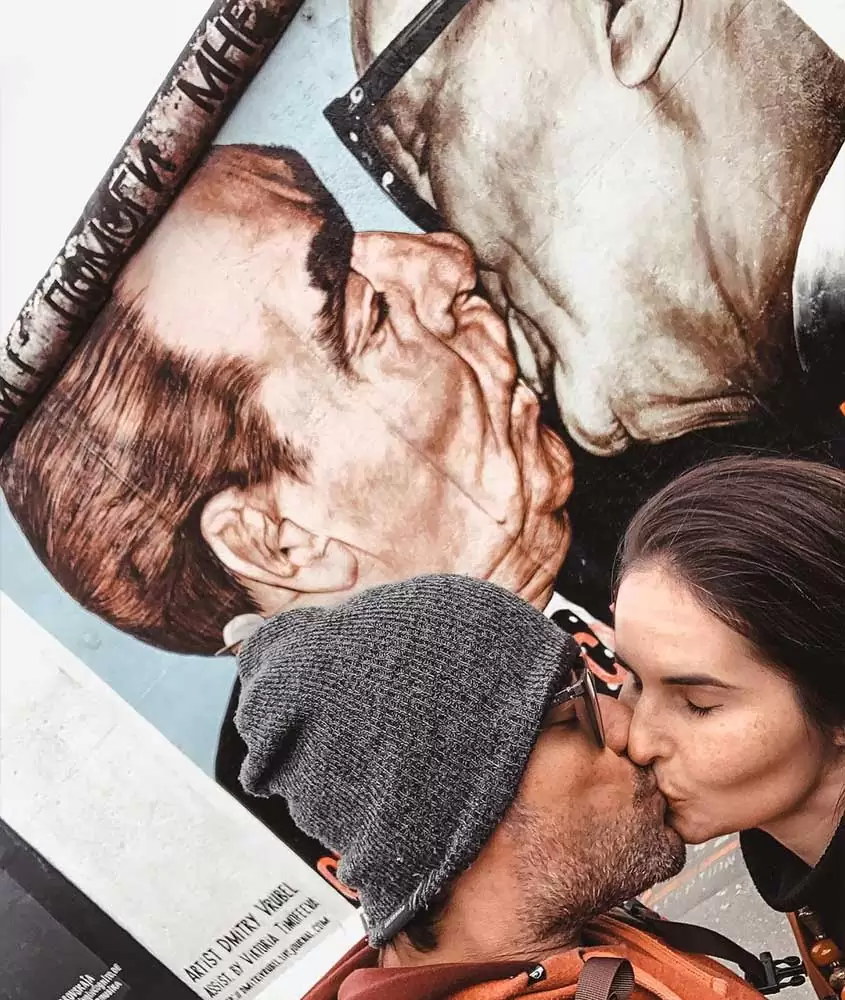 casal viajando o mundo, vagner, de touca cinza e casaco laranja e bárbara, de cabelo solto, se beijam, durante o dia, em frente a pintura em muro de berlim, que exibe beijo entre dois políticos