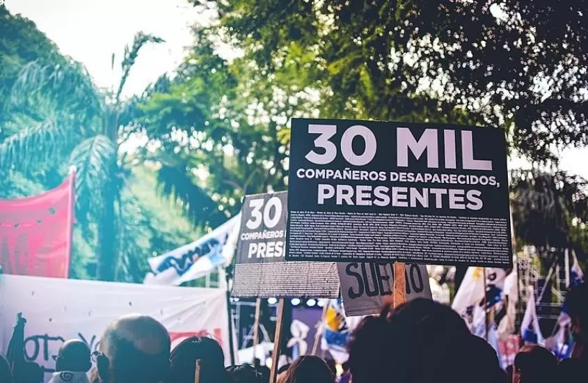 durante o dia, argentinos protestam segurando cartaz preto escrito em branco '30 mil compañeros desaprecidos, presentes'