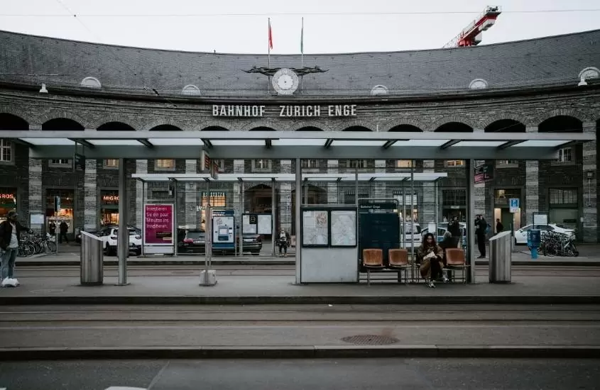 durante o dia, estação de ônibus e ao fundo, construção de concreto cinza onde há escrito 'bahnhof zurich enge'