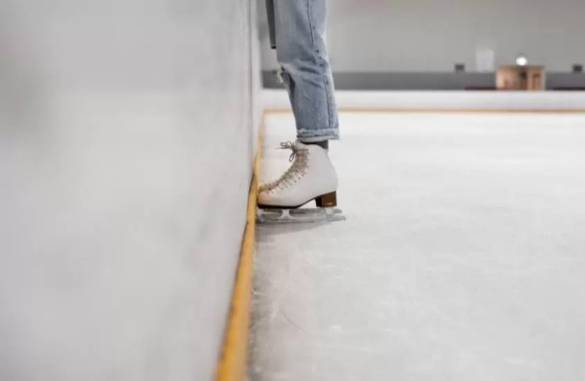 perna de pessoa, vestindo calça jeans e calçando patins, pisando em pista de patinação no gelo
