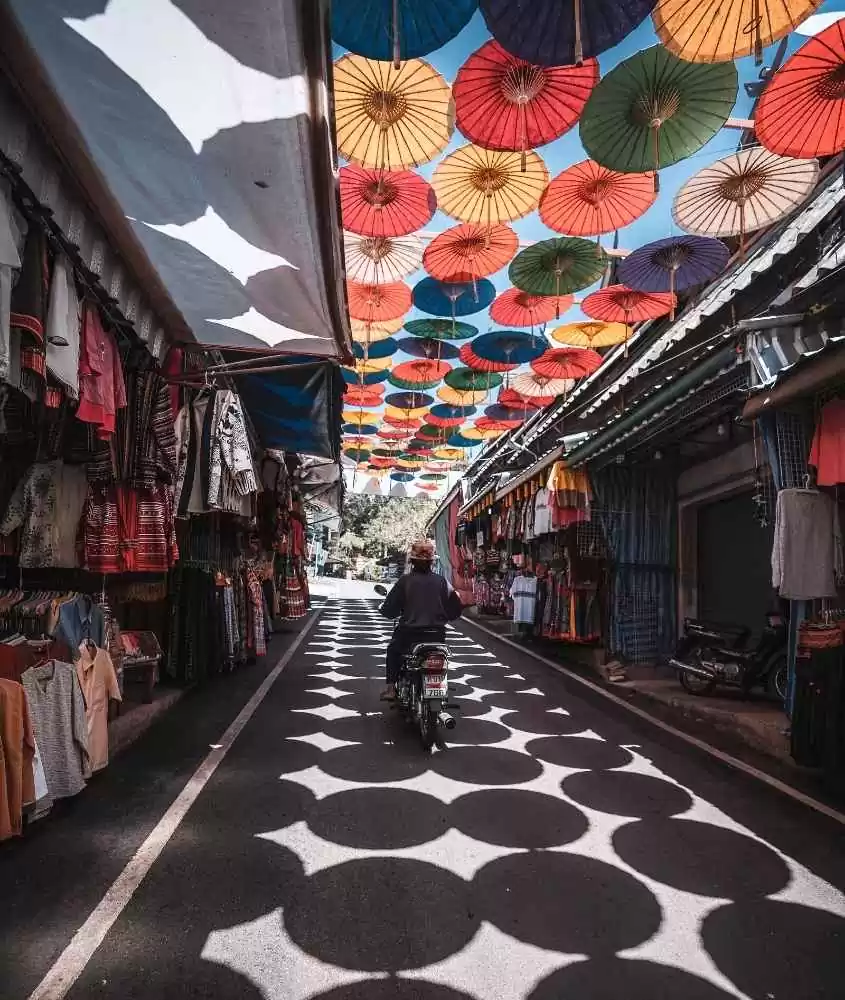 durante o dia, pessoa passando de moto entre barracas de roupas, em rua coberta por guarda-chuvas coloridos