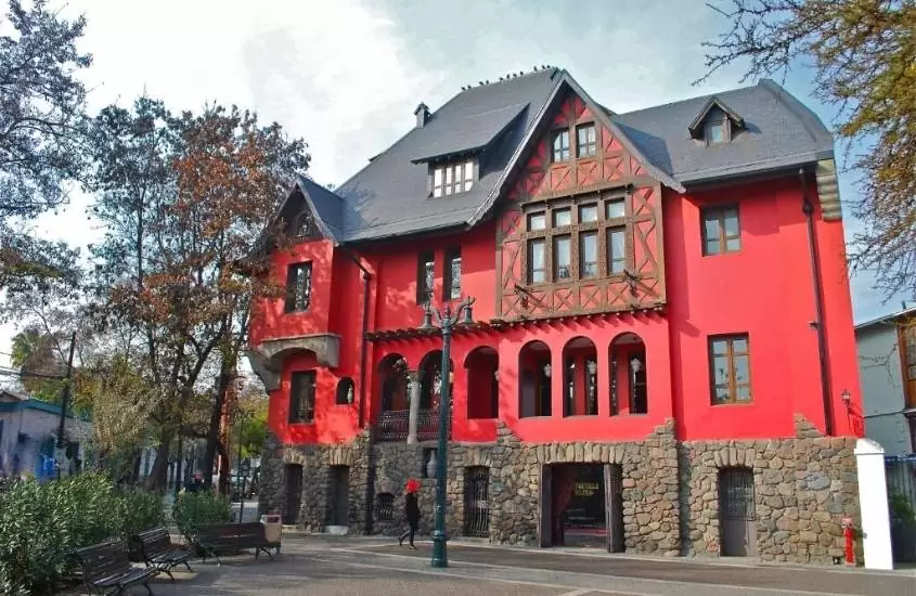 durante o dia, árvores em frente a casa 4 andares vermelha com telhado preto, onde funciona um dos hotéis em santiago do chile