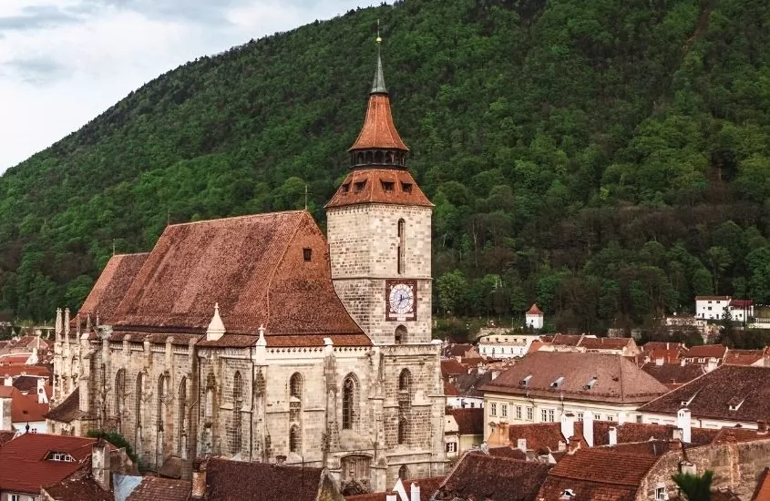 durante o dia, casas e igreja com telhado marrom e montanha ao fundo em brasov, destino para um roteiro romênia