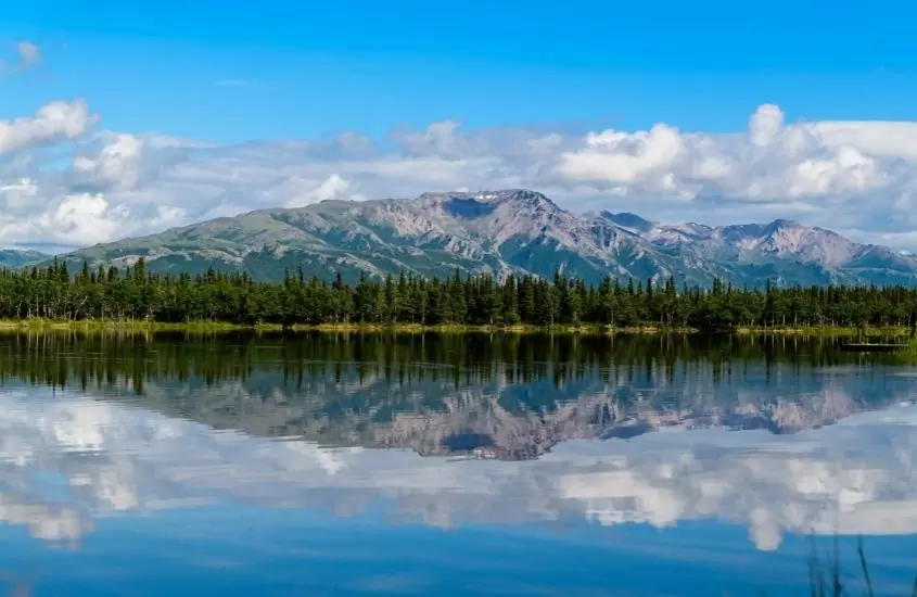durante o dia, grande lago cercado por árvores e, ao fundo, montanhas em alasca, que segundo curiosidade dos estados unidos foi comprado da rússia