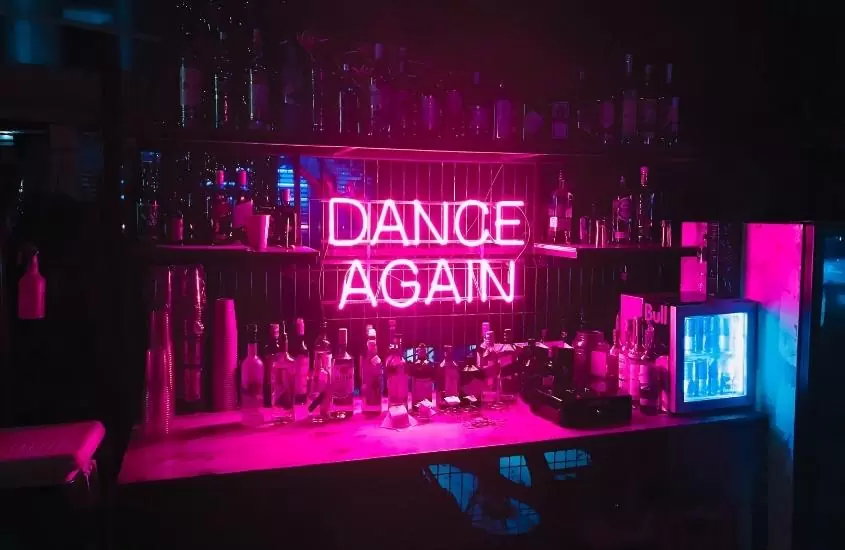 garrafas de bebidas em balcão de bar, onde há letreiro de led rosa escrito 'dance again'
