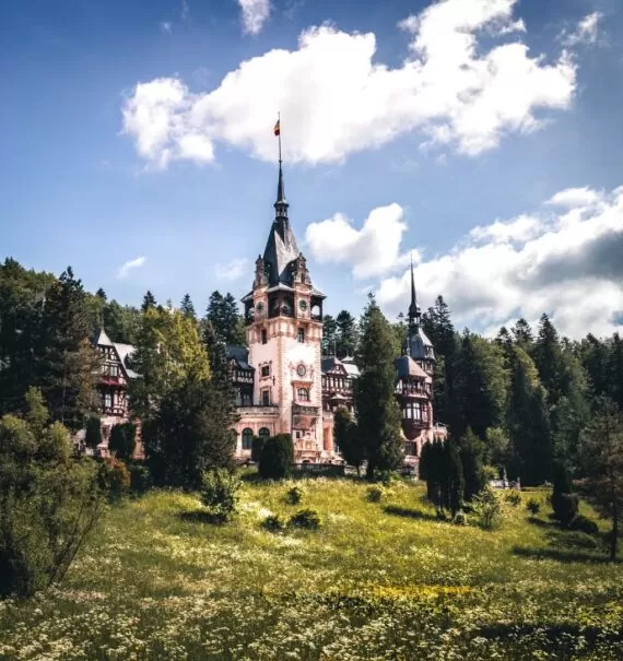 durante o dia, construção branca com telhado cinza, onde funciona um dos castelos da romênia, rodeado por diversas árvores verdes