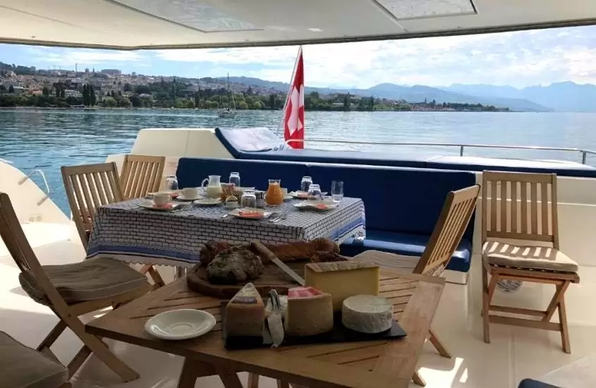 durante o dia, deck de barco, onde estão cadeiras ao redor de mesas quadradas, onde estão servidos sucos, queijos e frutas
