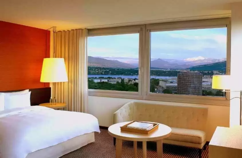 cama de casal, sofá e mesa redonda de centro, em suíte de hotel em genebra com ampla janela com vista para a cidade, durante o dia