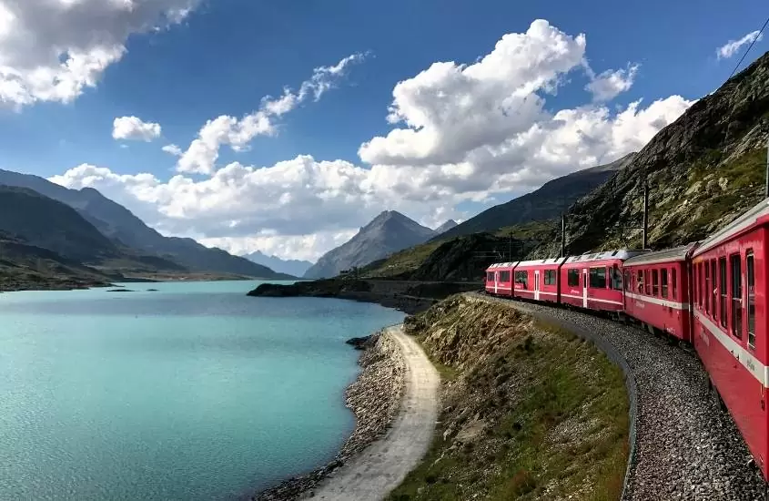 durante o dia, trem vermelho passando em frente a lago azul