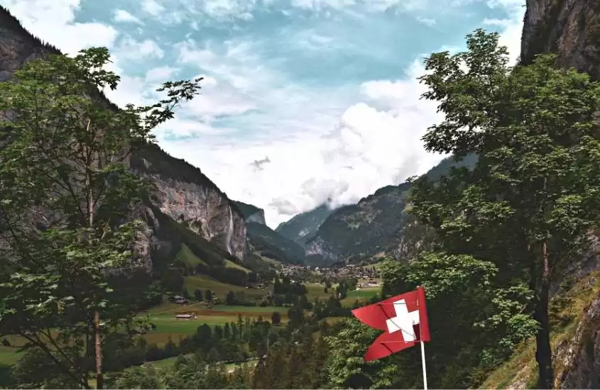 durante o dia, bandeira vermelha com cruz branca, ao fundo, diversas árvores e montanhas