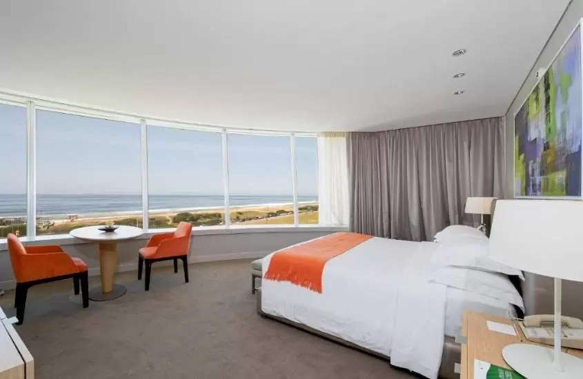 durante o dia, cama de casal e duas cadeiras ao redor de mesa redonda, em suite com ampla janela com vista para o mar