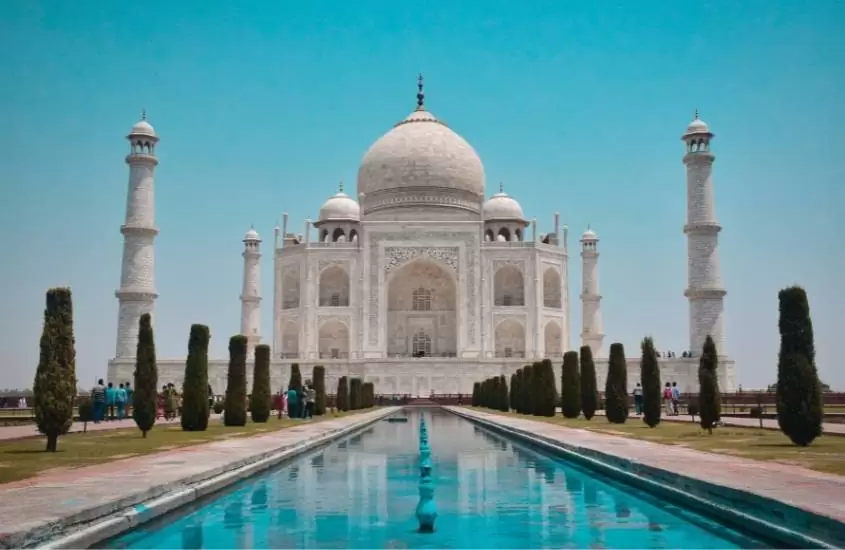 durante o dia, grande piscina azul em frente a mausoléu de mármore branco que é um dos pontos turísticos da Índia