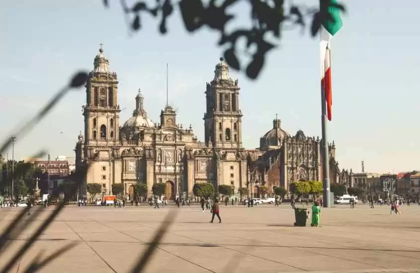 durante o dia, pessoas caminhando em praça onde há hasteada bandeira do méxico, ao fundo, grande igreja