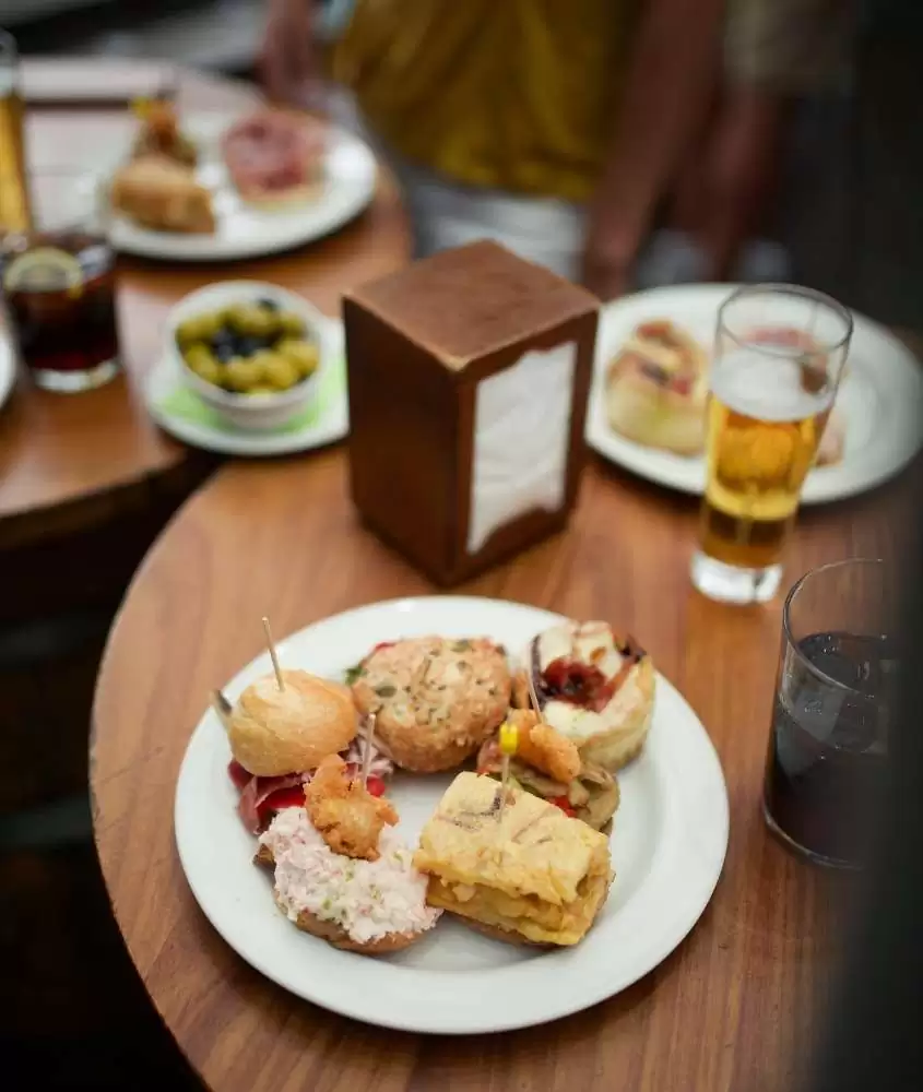vista de cima de prato branco, cheio de tapas, e copo de cerveja e refrigerante em cima de mesa de madeira